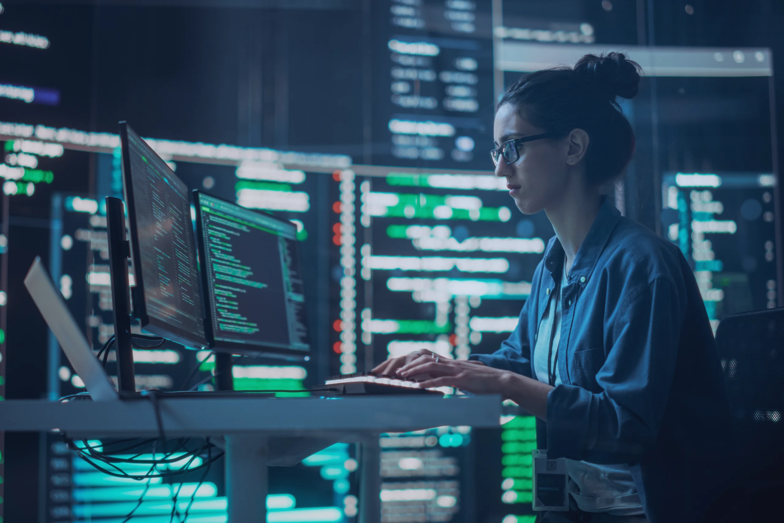 A woman with glasses and her hair in a bun types at a multi-monitor workstation displaying code, in a dark room filled with