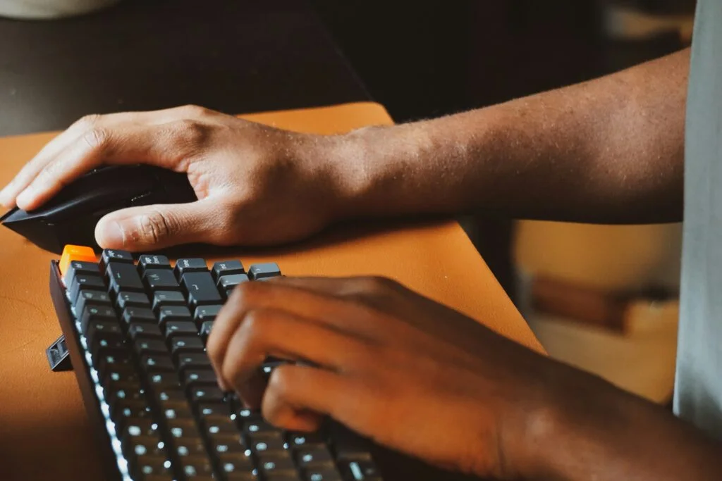 Two hands using a black computer mouse and typing on a black keyboard with one orange keycap on an orange surface.