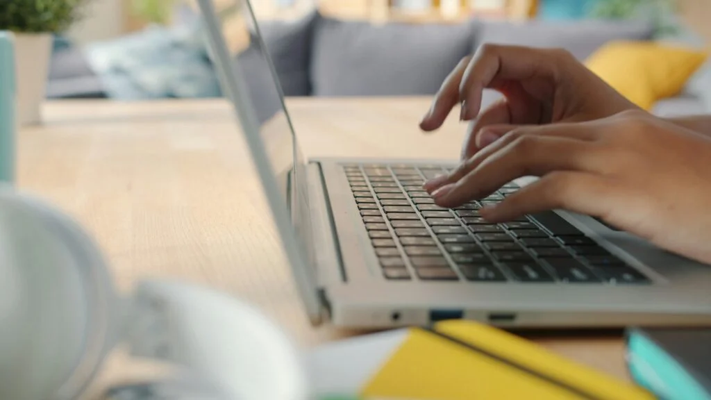 Hands typing on a silver laptop keyboard on a wooden table, with a blurred sofa and cushions in the background.